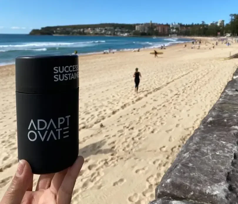 A photograph shows a black cylindrical container labeled 'ADAPT OVATE' and 'SUCCESS SUSTAIN' held up against the backdrop of Manly Beach in Sydney, Australia. The beach scene features golden sand, blu