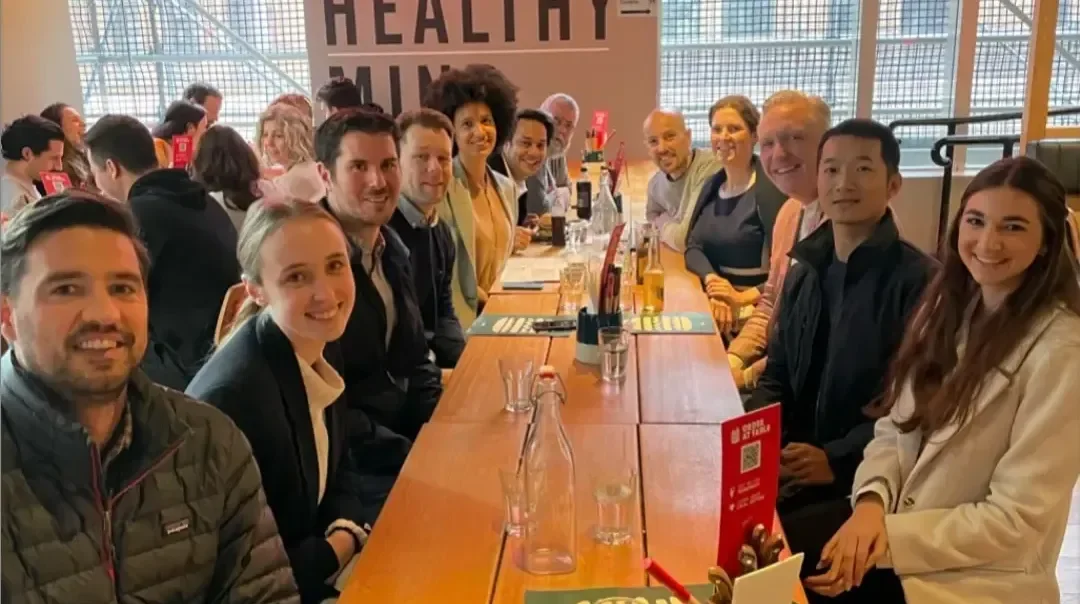 A group of professionals gathered at a restaurant or casual dining establishment with 'HEALTHY BURGERS HEALTHY' signage prominently displayed on the wall behind them. The scene shows a long wooden tab