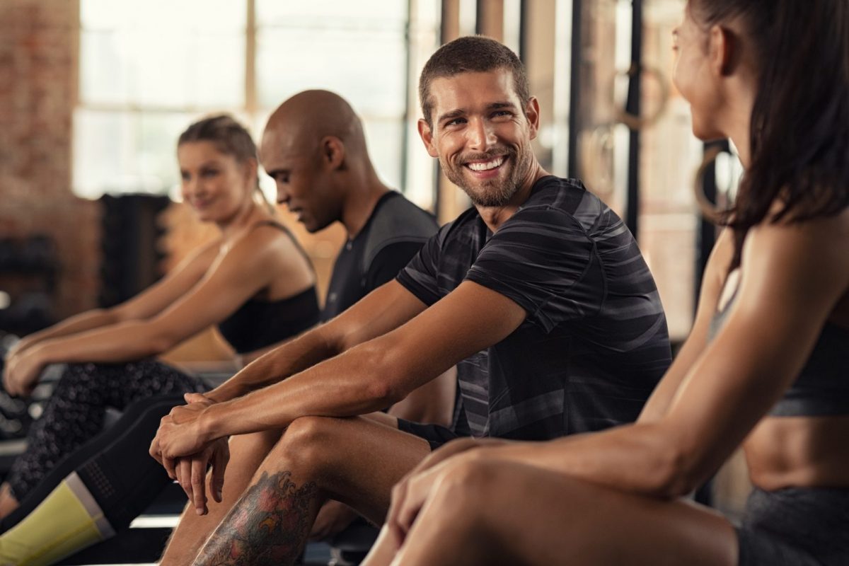 A candid gym scene showing people taking a break during a workout session. The image is shot in a fitness studio with natural lighting through large windows. The individuals are sitting and appear to